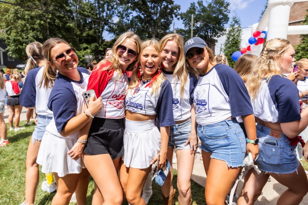 A group of young women dressed in casual summer attire pose together at an outdoor event. They are wearing matching t-shirts with 'Chi Omega' on them, celebrating with balloons and a festive atmosphere in the background.