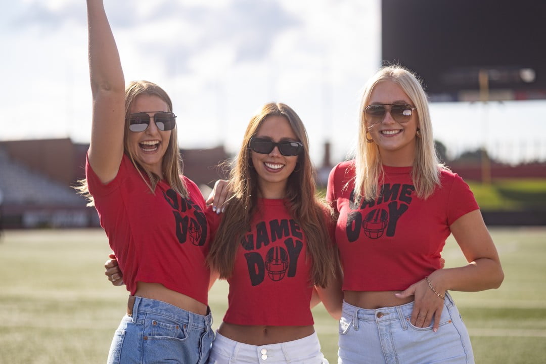 Three young women are standing in a stadium, wearing bright red crop tops with 'Game Day' printed on them. They are wearing sunglasses and denim shorts, with one raising her arm in a cheer, capturing the excitement of a sports event day.