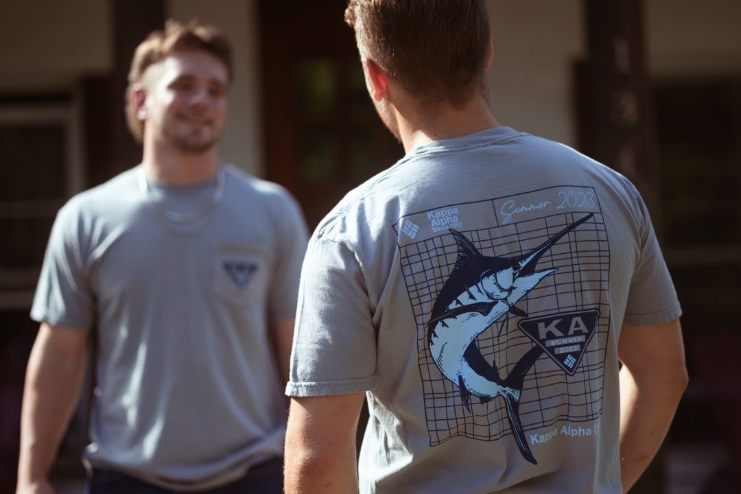 A young man is seen from the back wearing a light grey t-shirt with a large print of a shark and 'Kappa Alpha Order' on it. In the foreground, another young man is facing the camera, slightly out of focus, creating a candid atmosphere.