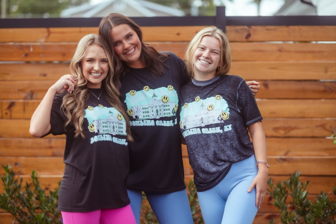 Three young women wearing matching dark t-shirts with a colorful graphic print are posing with smiles. They appear to be in a casual outdoor setting with a wooden fence and greenery in the background.