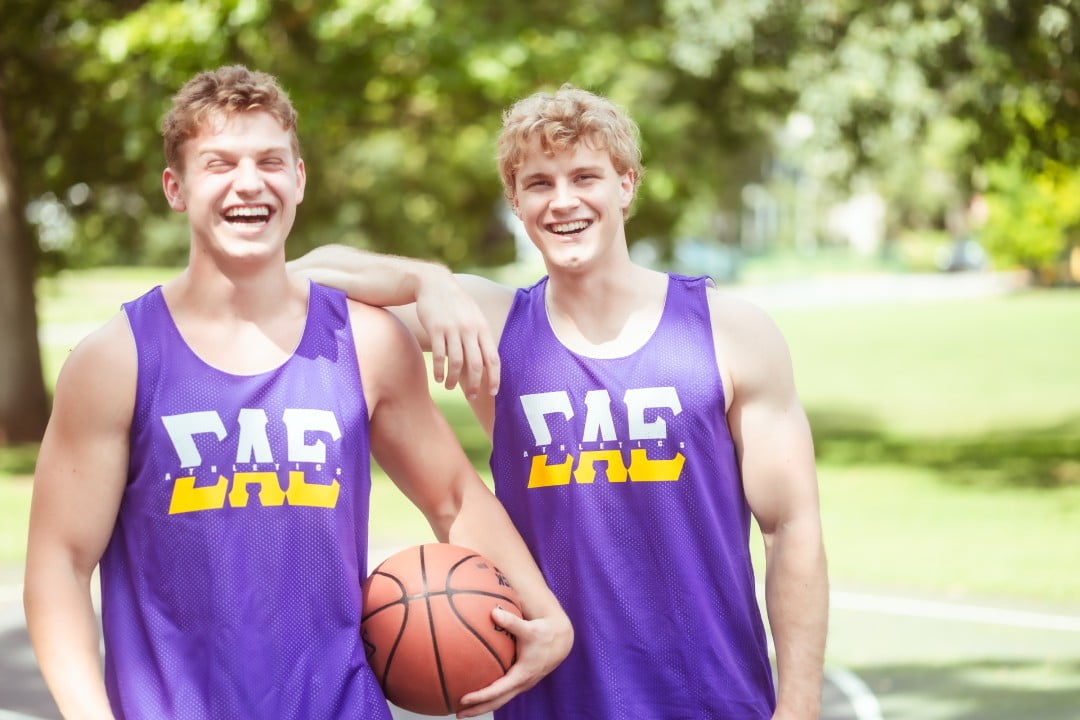 Two young men wearing purple and gold basketball tank tops with 'ΣΑΕ' printed on them are standing on a basketball court, sharing a laugh. One is holding a basketball. They are casually dressed for a game or practice, and trees can be seen in the soft-focus background.