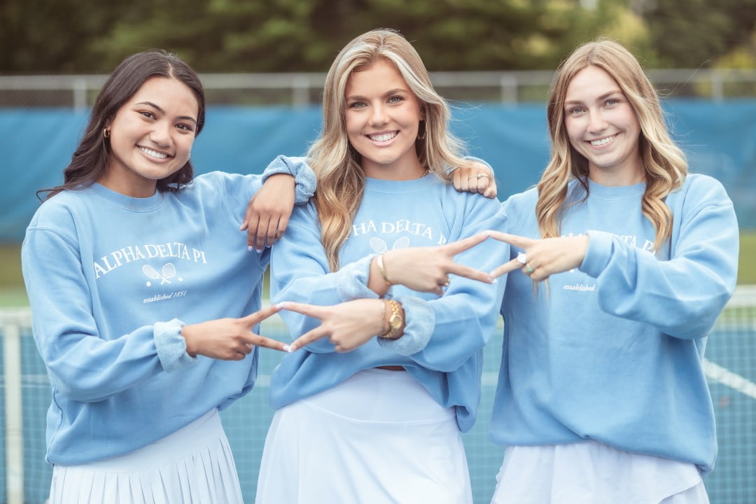 Three young women in matching light blue sweatshirts with 'Alpha Delta Pi' printed on them are standing on a tennis court. They are posing with their hands making a sign that appears to be a symbol associated with their organization. The day is overcast, and they are all smiling at the camera.