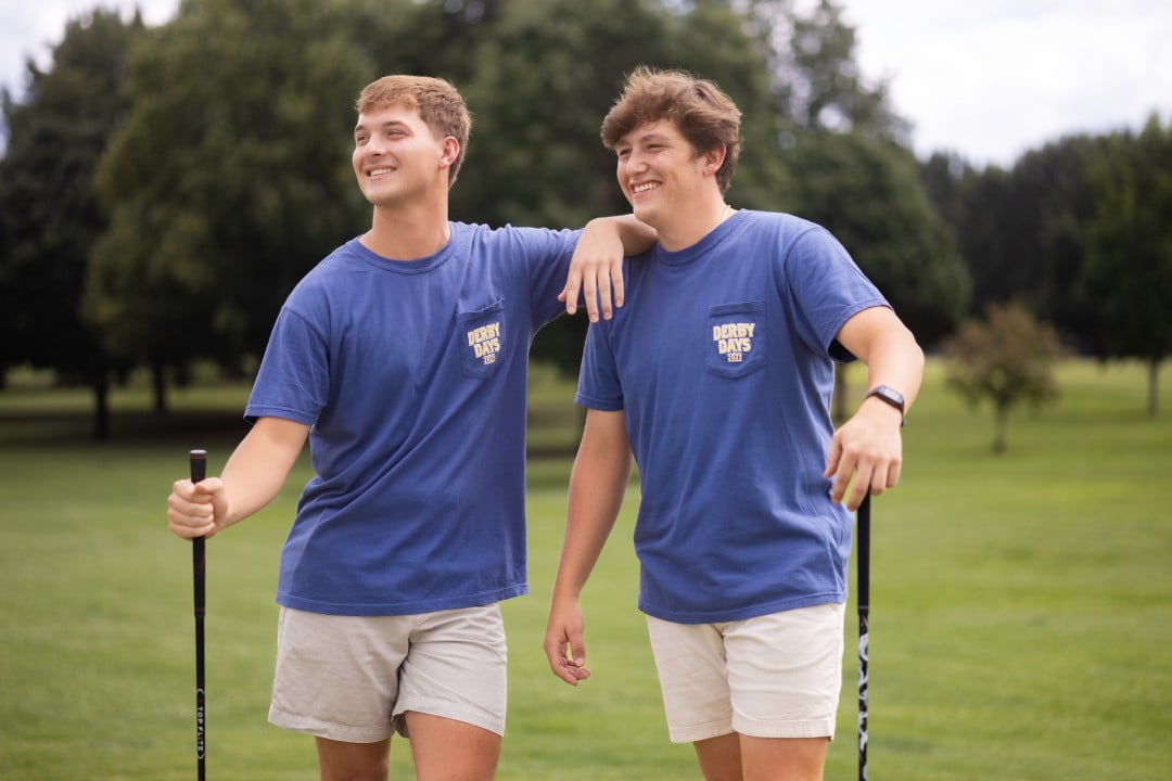 Two young men wearing matching blue t-shirts with 'Derby Days' printed on them are standing on a golf course. They are both holding golf clubs, smiling, and seem to be enjoying their game. The setting is outdoors with trees and a cloudy sky in the background.